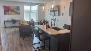 A modern kitchen with a central island, bar stools, pendant lighting, and a dining area with a sofa and chairs. - Gated 2BR in Greenville w/Vibes (Greenville)