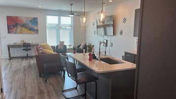 A modern kitchen with a central island, bar stools, pendant lighting, and a dining area with a sofa and chairs.