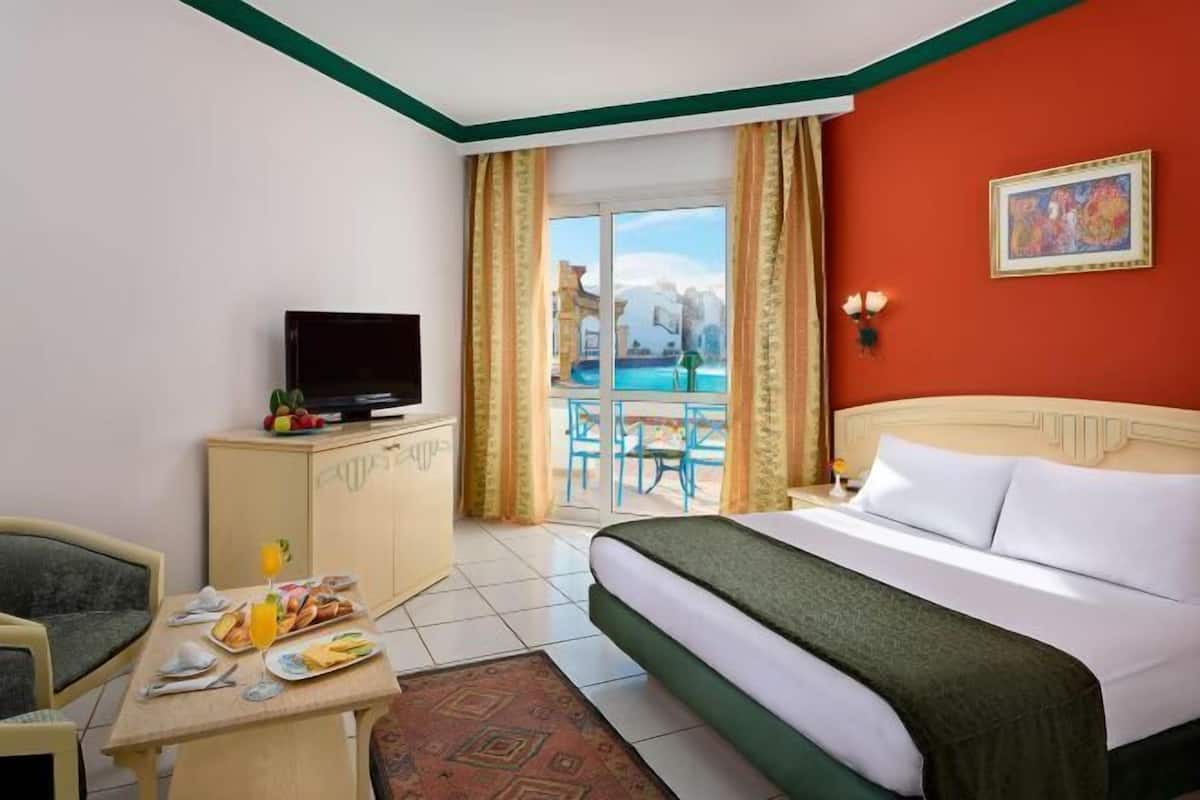 A hotel room with a bed, a desk with breakfast, a TV, and a view of a pool and building.