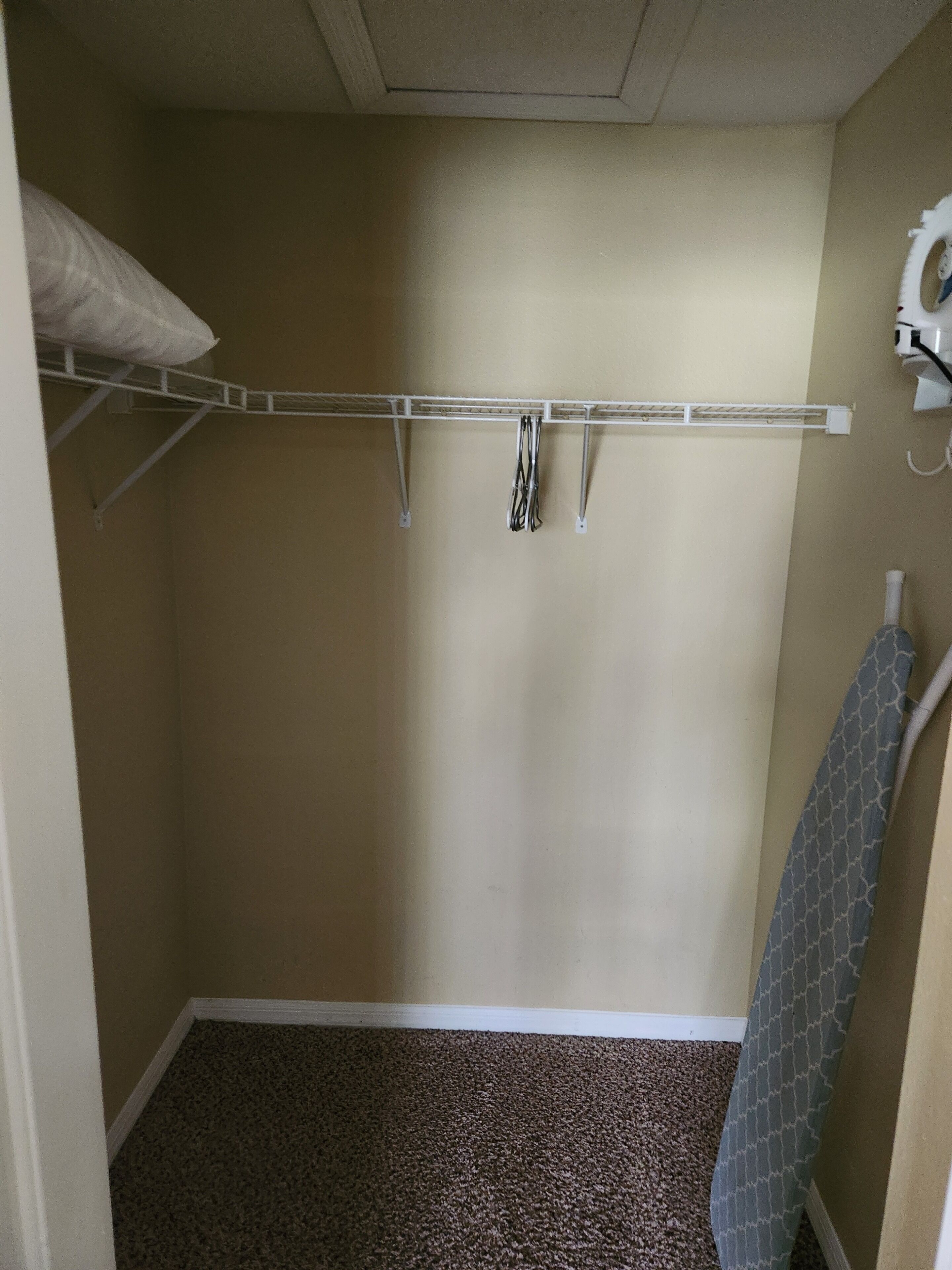 Een kleine, smalle kamer met een beige muur, een wit plafond en een bruin tapijt. Er staat een wit plankje met een wit kussen erop, en een witte hangrail met kleding.