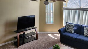 A living room with a ceiling fan, a television on a stand, a sofa with patterned cushions, and a large window with blinds. - 3-bedroom condo in welcoming Kissimmee with WiFi, AC and Gated community. (Kissimmee)