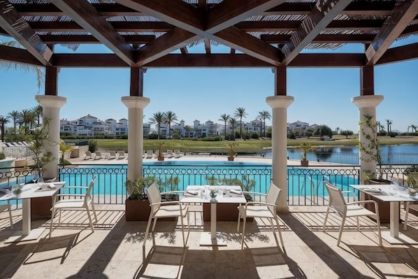 A covered outdoor area with a pool, tables, and chairs, overlooking a body of water and buildings in the distance. - DoubleTree by Hilton La Torre Golf & Spa Resort (Murcia)