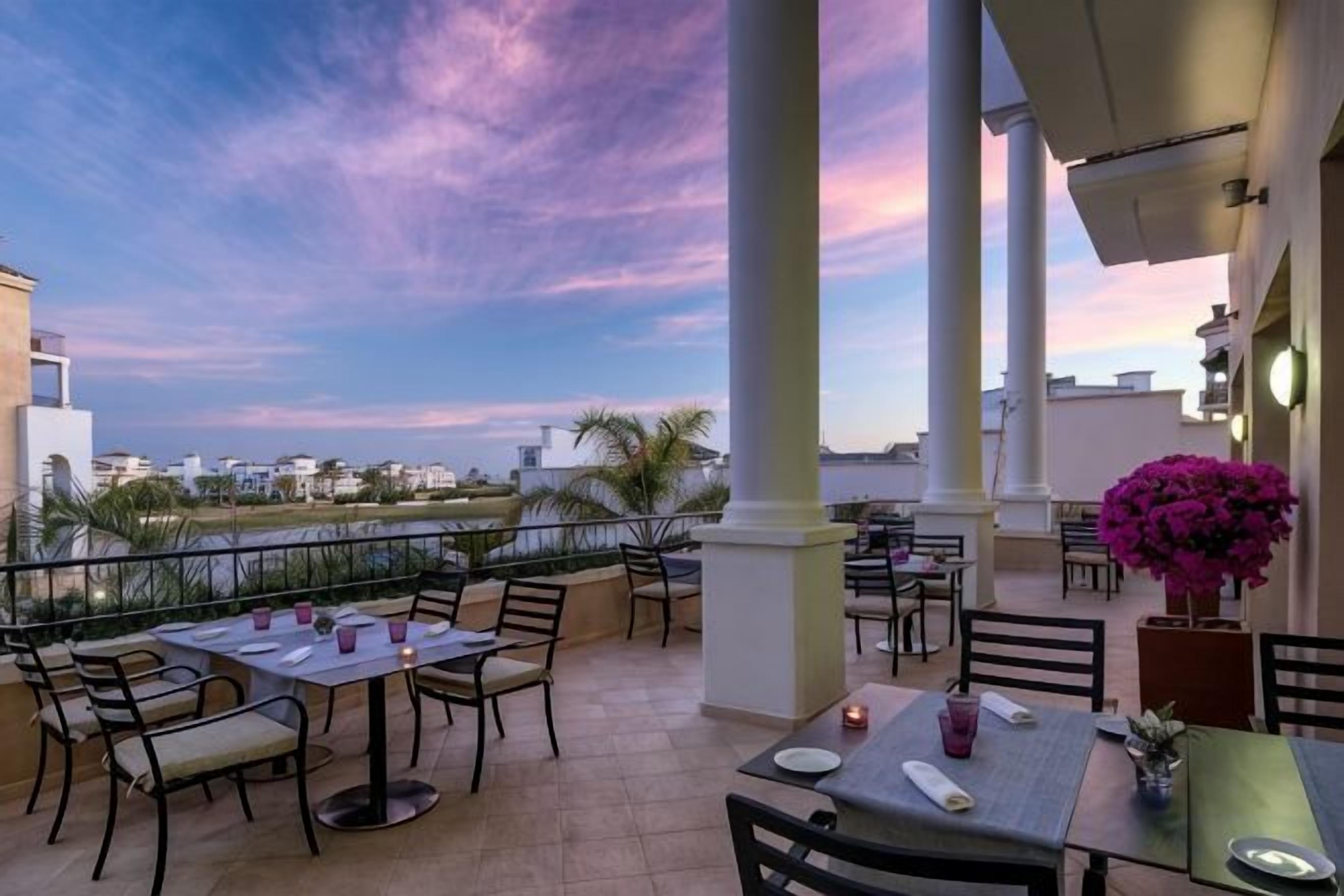 An outdoor terrace with tables and chairs, a view of the sea, and a building with columns.