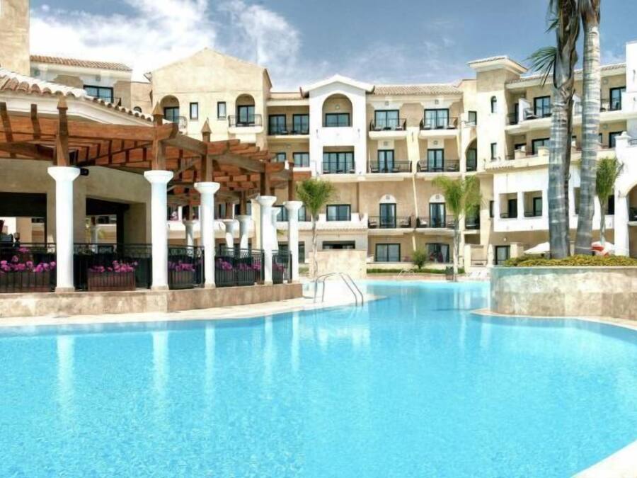 A swimming pool surrounded by multi-story buildings with balconies, a covered area with columns, and palm trees.