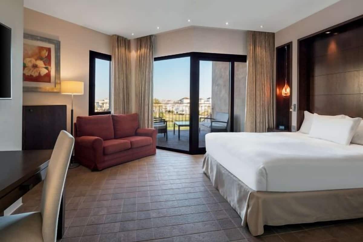 A hotel room with a bed, a desk, a chair, a red sofa, and a large window with a view.