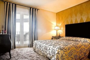 Una camera d'albergo con un letto, un comodino, una lampada, una cassettiera e vista attraverso una finestra con tende.