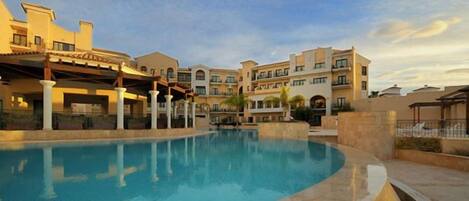 A swimming pool with a curved edge, surrounded by buildings with multiple balconies and a covered area with columns.