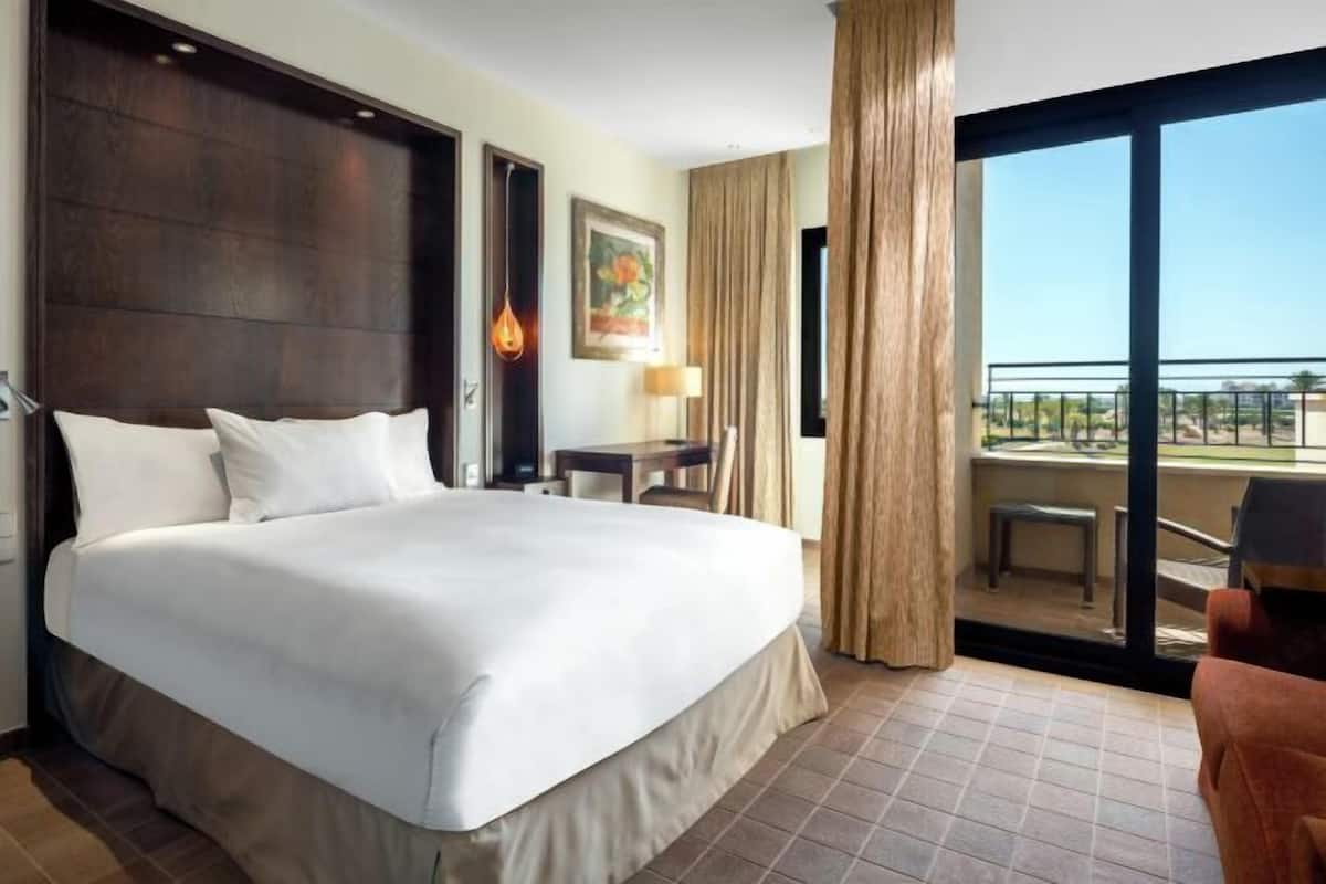 A hotel room with a large bed, a desk, a chair, a balcony with a view, and a painting on the wall.