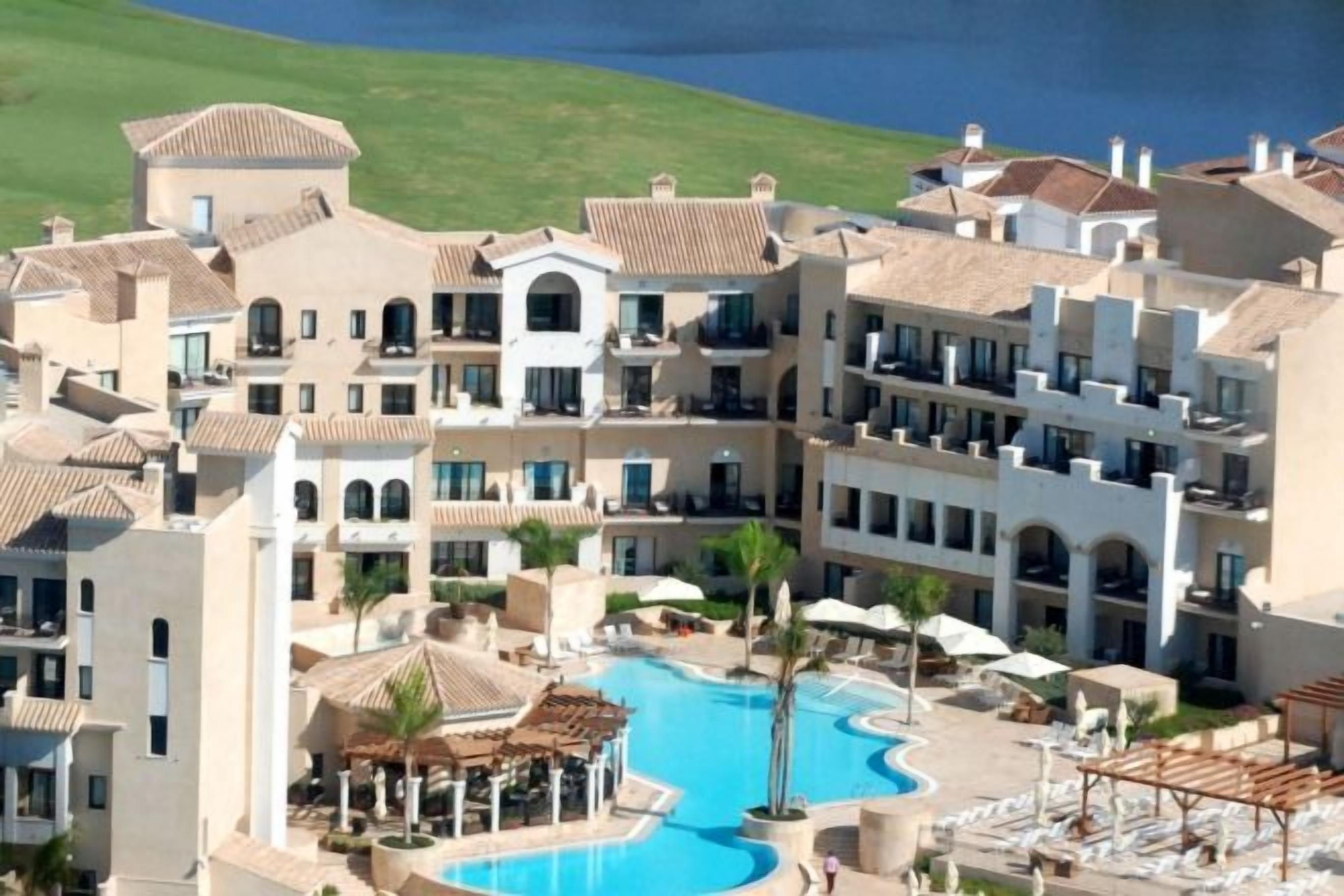 Un resort con varias edificaciones, una piscina central y vista al mar.