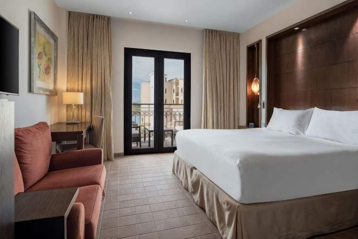 A hotel room with a bed, a desk, a chair, a TV, and a balcony with curtains.