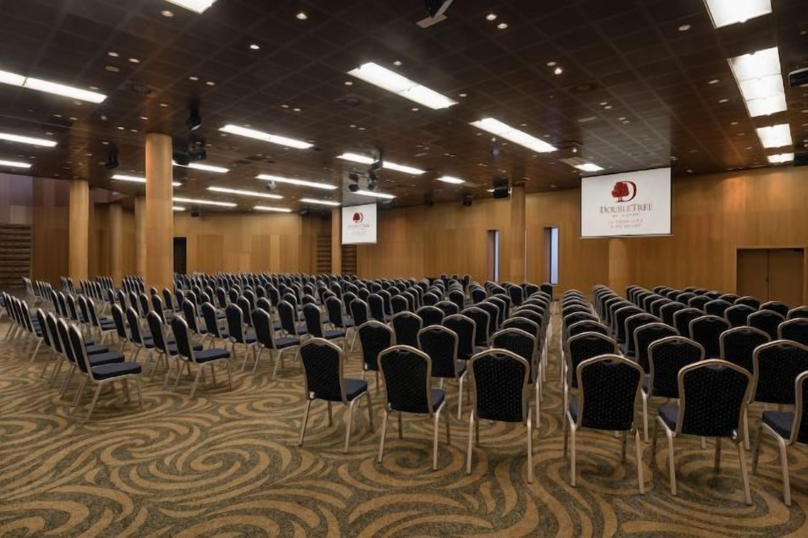 Een congreszaal met rijen stoelen, een podium en een spandoek.