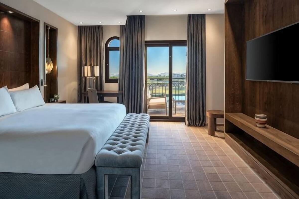 A hotel room with a large bed, a bench, a TV, a desk, and a balcony with a view.