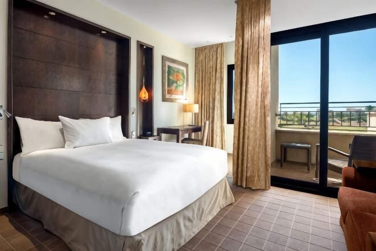 A hotel room with a large bed, a desk, a chair, a balcony with a view, and a painting on the wall.
