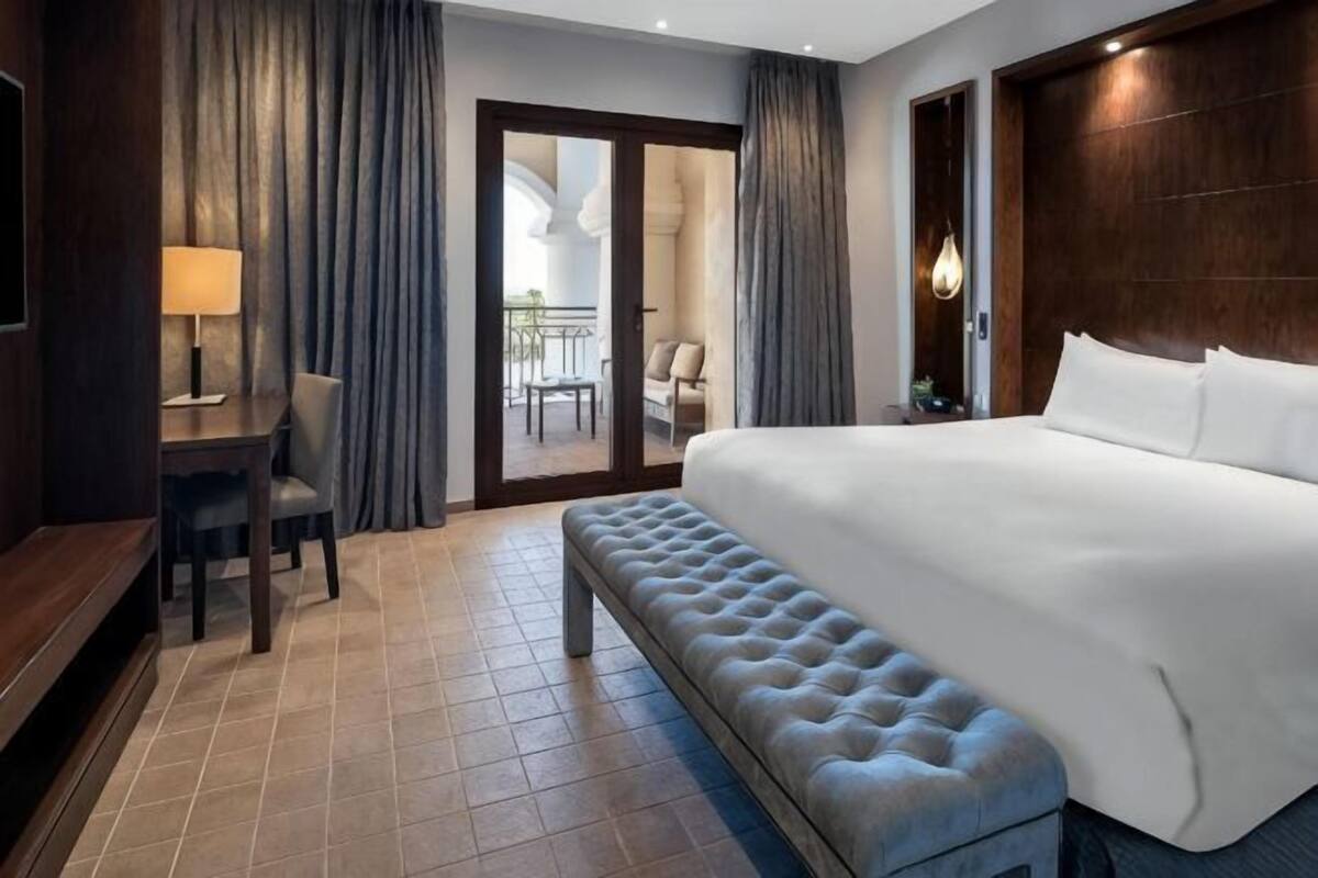 A hotel room with a large bed, a bench, a desk with a lamp, and a view of the outside balcony.