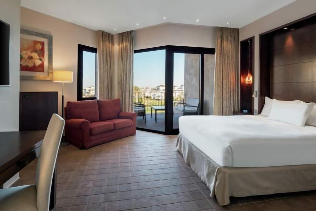 A hotel room with a bed, a red sofa, a desk, and a large window with curtains.