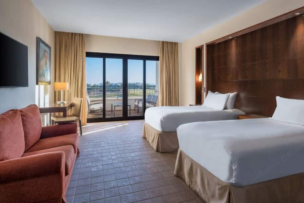 A hotel room with two beds, a sofa, a TV, a desk, and a large window with a view.