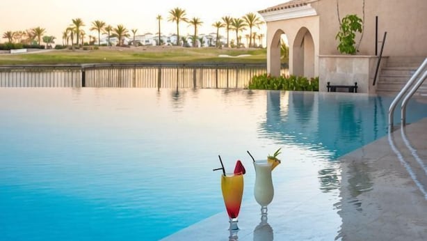A swimming pool with two drinks on the edge, a palm tree-lined background, and a building with arches. - DoubleTree by Hilton La Torre Golf & Spa Resort (Murcia)