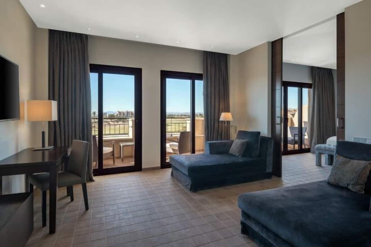 A hotel room with a sofa, a chair, a TV, a table, and a balcony with a view.