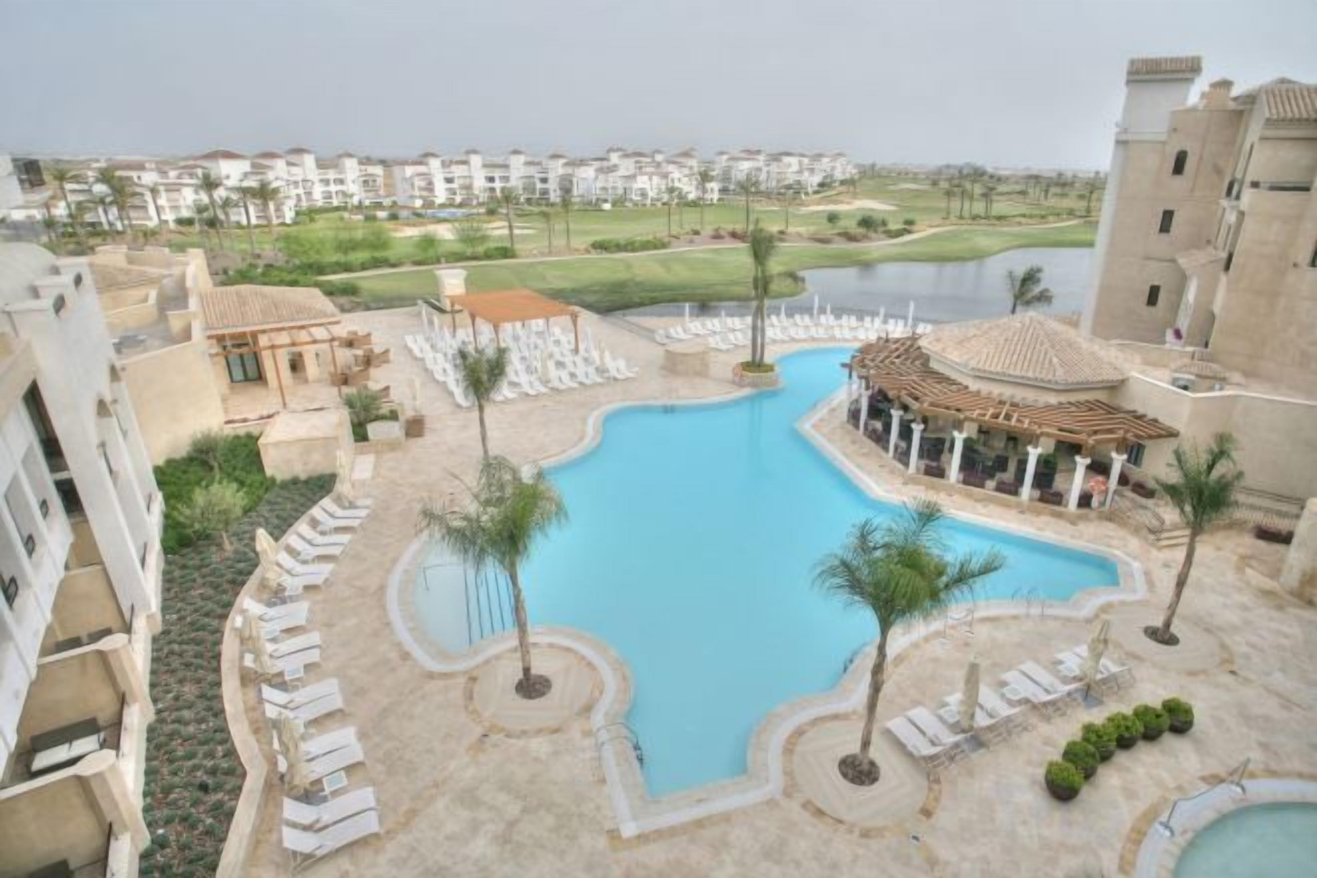 Una piscina con sillones de descanso y una pérgola, rodeada de una zona turística con edificios y un campo de golf al fondo.