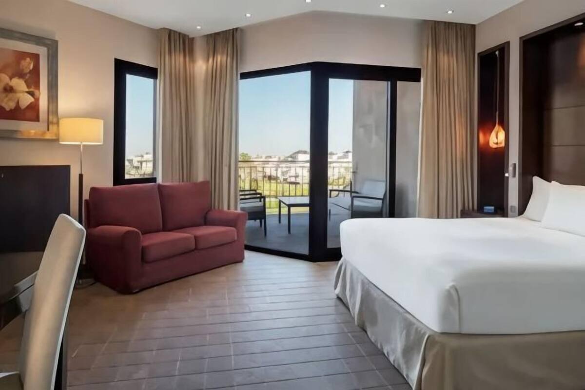 A hotel room with a bed, a red sofa, a desk, and a balcony with a view.