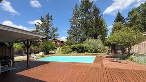 A wooden deck with a swimming pool, lounge chairs, and a covered patio area.