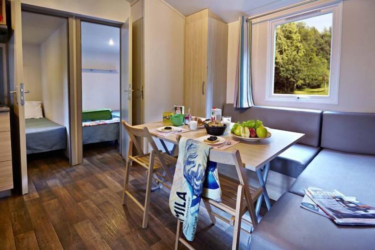Quarto compacto com mesa de jantar, cadeiras, um banco e janela com vista para árvores.