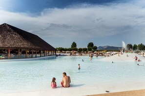 Een groot buitenzwembad met een paviljoen met rieten dak, omringd door parasols en bomen.