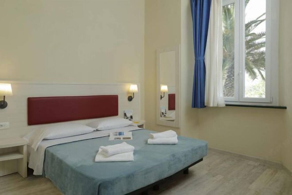 Quarto de hotel com cama, mesas de cabeceira, luminárias, janela com cortinas e vista para palmeiras.