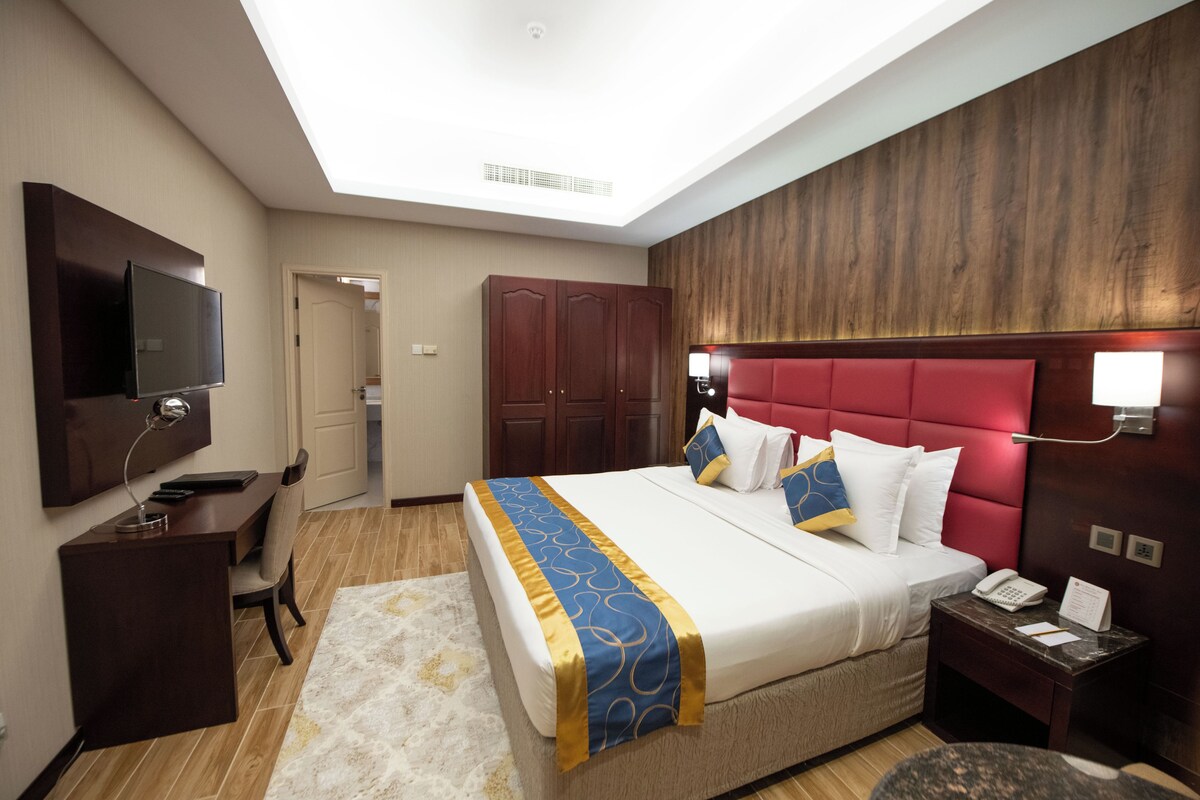 A hotel room with a large bed, a desk with a chair, a television, and a wardrobe.