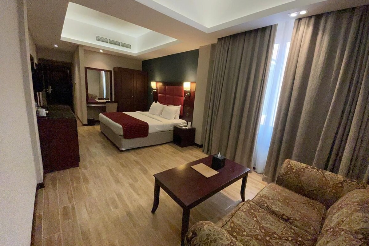 A hotel room with a bed, a sofa, a table, a mirror, and a window with curtains.