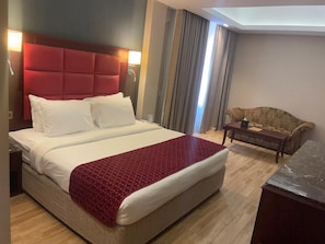A hotel room with a large bed, a red headboard, a sofa, a small table, and a view of the outside. - Ramee Guestline Hotel (Muscat)