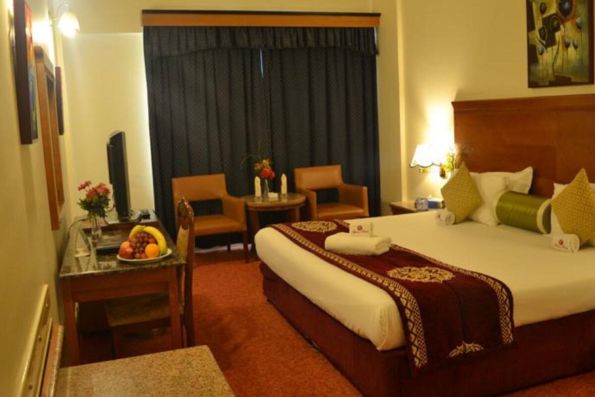 Una habitación de hotel con una cama grande, un escritorio con fruteros, dos sillas, una mesita y un cuadro en la pared.