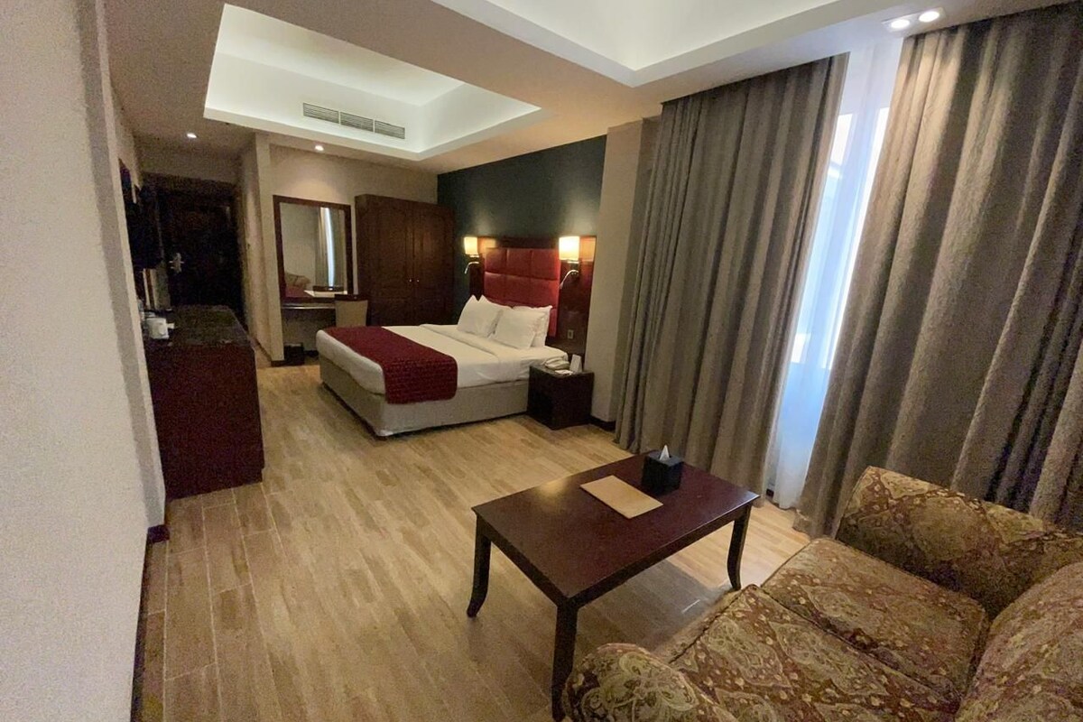 Ein Hotelzimmer mit Bett, einer Sitzecke, einem Couchtisch, einem Schreibtisch und einem großen Fenster mit Vorhängen.