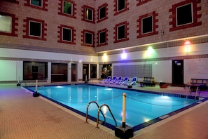 An indoor swimming pool with a tiled floor, poolside seating, and a brick wall with windows. - Ramee Guestline Hotel (Muscat)
