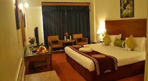 A hotel room with a large bed, a desk with a fruit bowl, two chairs, a small table, and a painting on the wall. - Ramee Guestline Hotel (Muscat)