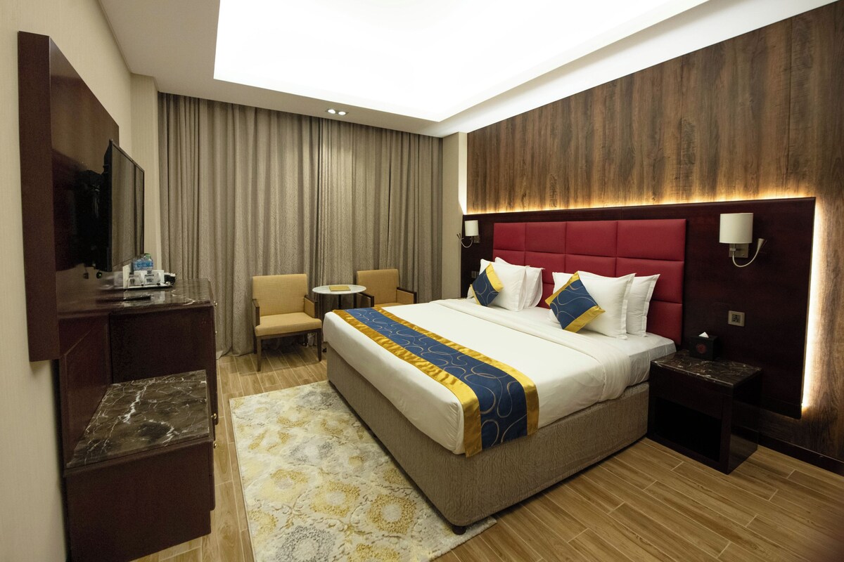 Habitación de hotel con una cama grande, un escritorio con televisor de pantalla plana, una silla, una mesita redonda y una mesita de noche con lámpara.