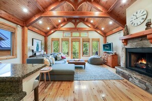 Un vaste salon doté d’une cheminée en pierre, de poutres apparentes en bois et d’une grande fenêtre donnant sur un paysage verdoyant.