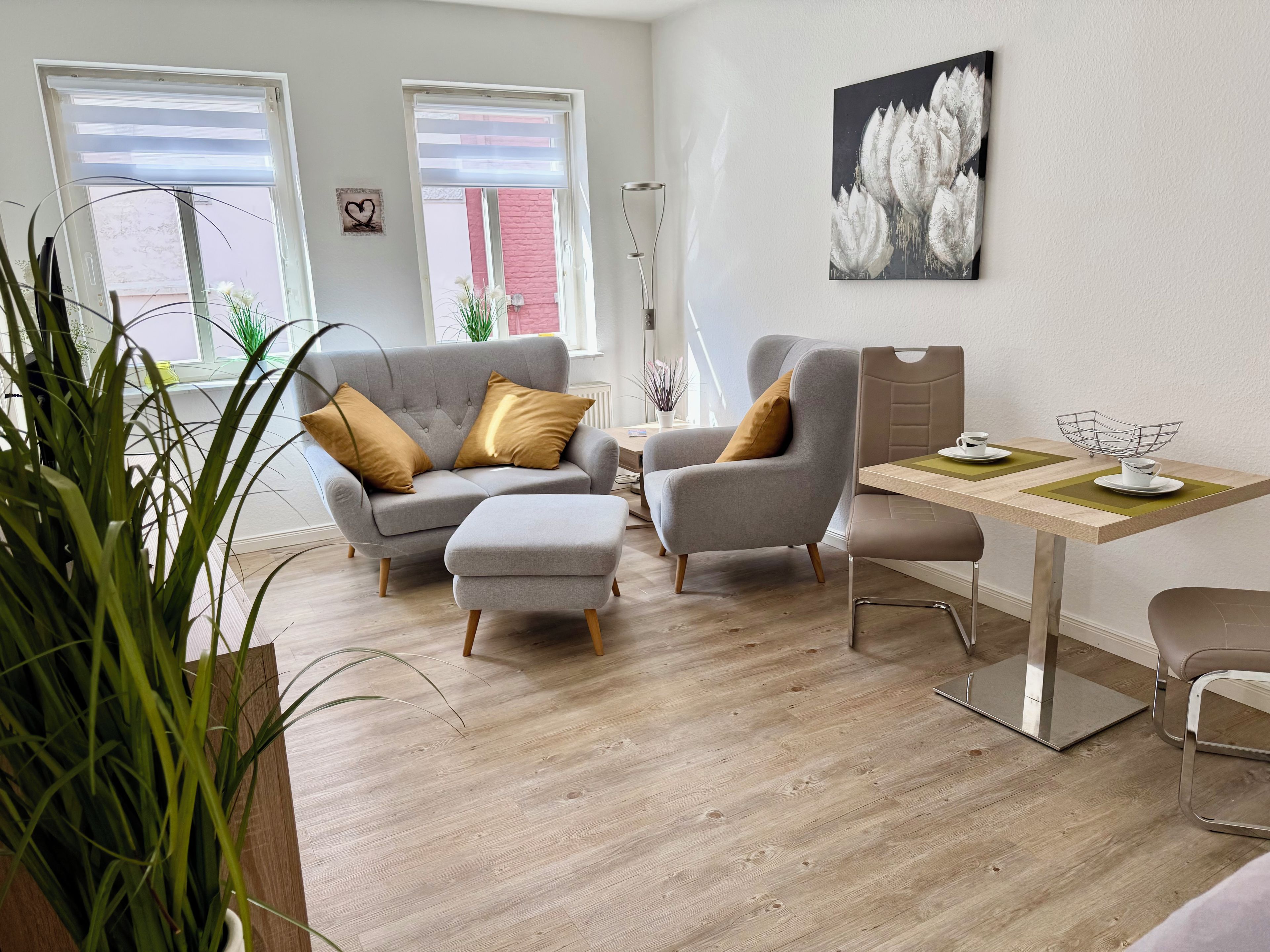 A modern living room with a grey sofa, ottoman, and armchair, a wooden dining table with chairs, and a large window with blinds.