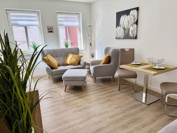 A modern living room with a grey sofa, ottoman, and armchair, a wooden dining table with chairs, and a large window with blinds.