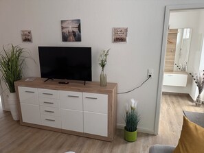 Una sala de estar con un televisor de pantalla plana sobre un mueble de madera, una planta en maceta y dos cuadros colgados en la pared.
