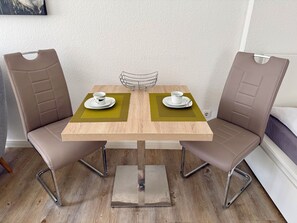 Une petite table de salle à manger carrée, accompagnée de deux chaises, chacune dotée d’un siège et d’un dossier rembourrés. La table a un cadre en métal et est posée sur un plancher en bois.