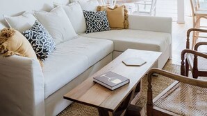 Ein Wohnzimmer mit einer Couch, einem Couchtisch und einem Sessel. Ein Gemälde hängt an der Wand und es gibt einen Blick ins Freie.