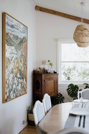 A dining area with a wooden table, chairs, a cabinet with decorative items, a hanging light fixture, and a large painting on the wall.