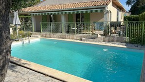 A swimming pool with a tiled deck, a covered seating area, and a building with a tiled roof and green shutters. - Quiet villa with pool close to the sea (Marsillargues)