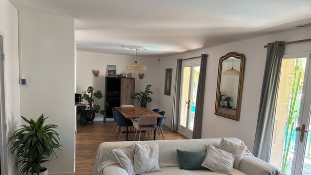 A living room with a sofa, coffee table, and dining area with chairs and a table. There is a large window with curtains, a mirror on the wall, and a potted plant. - Quiet villa with pool close to the sea (Marsillargues)