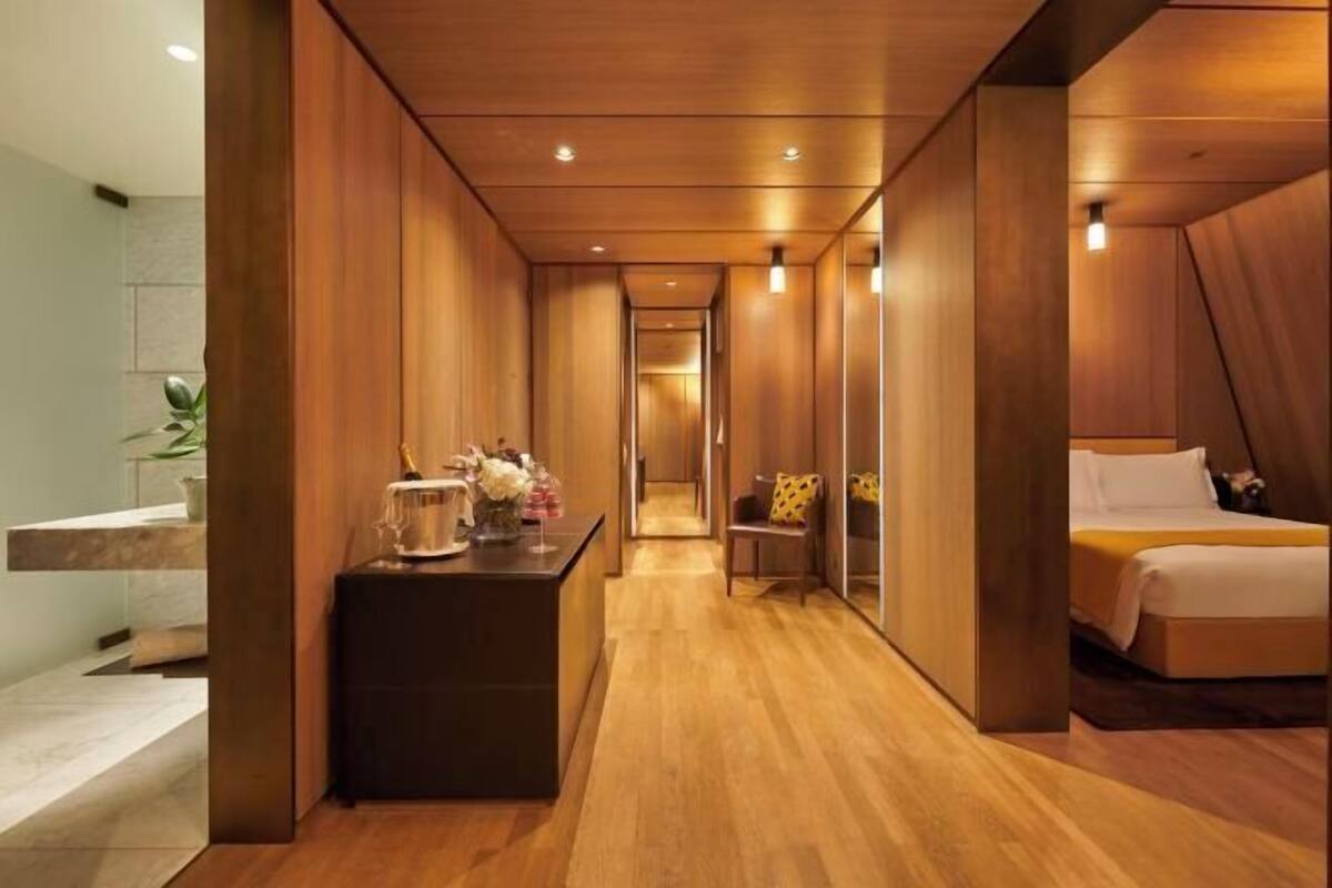 A modern hotel room with a wooden floor, a bed with white bedding, a small table with a vase of flowers, and a mirror on the wall.