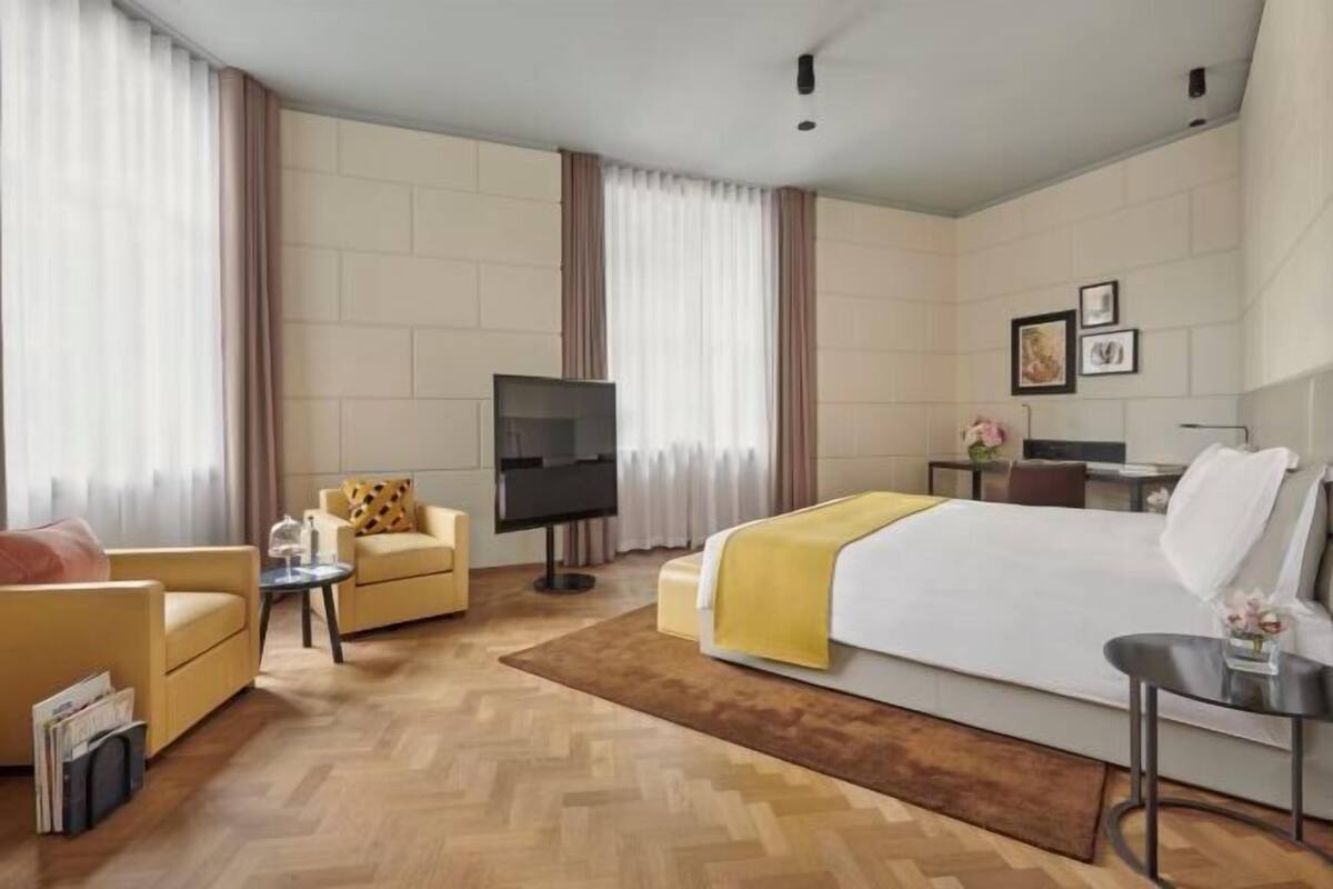 A hotel room with a bed, two armchairs, a small table, a TV, and a desk with a chair.