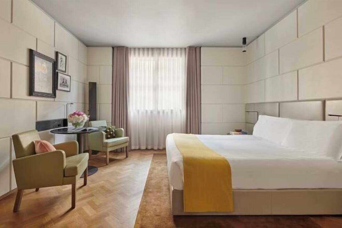 A hotel room with a large bed, a desk with a chair, a small table with a vase of flowers, and a window with curtains.