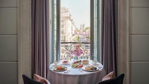 Una mesa de comedor con platos de comida, un florero y vistas de edificios a través de una ventana con cortinas.