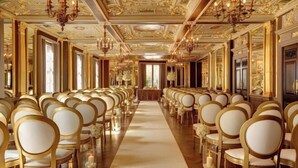Un gran salón con hileras de sillas doradas y un pasillo central, candelabros ornamentales y detallados adornos en el techo.